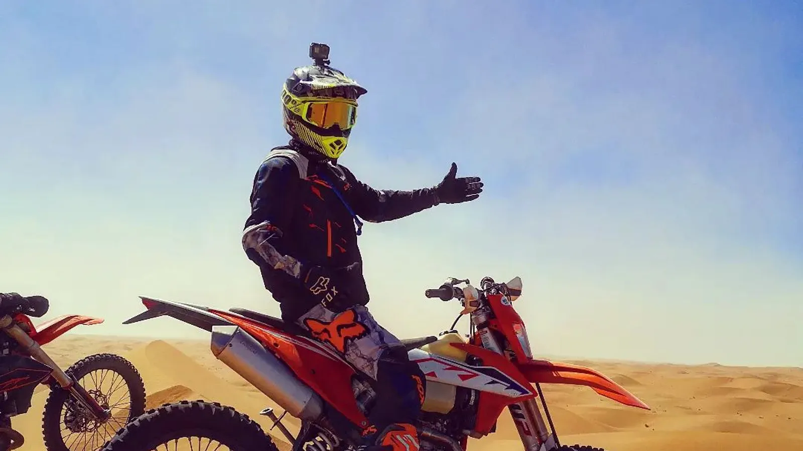 Pilote enduro détendu, geste de bienvenue, à côté de sa moto dans les dunes