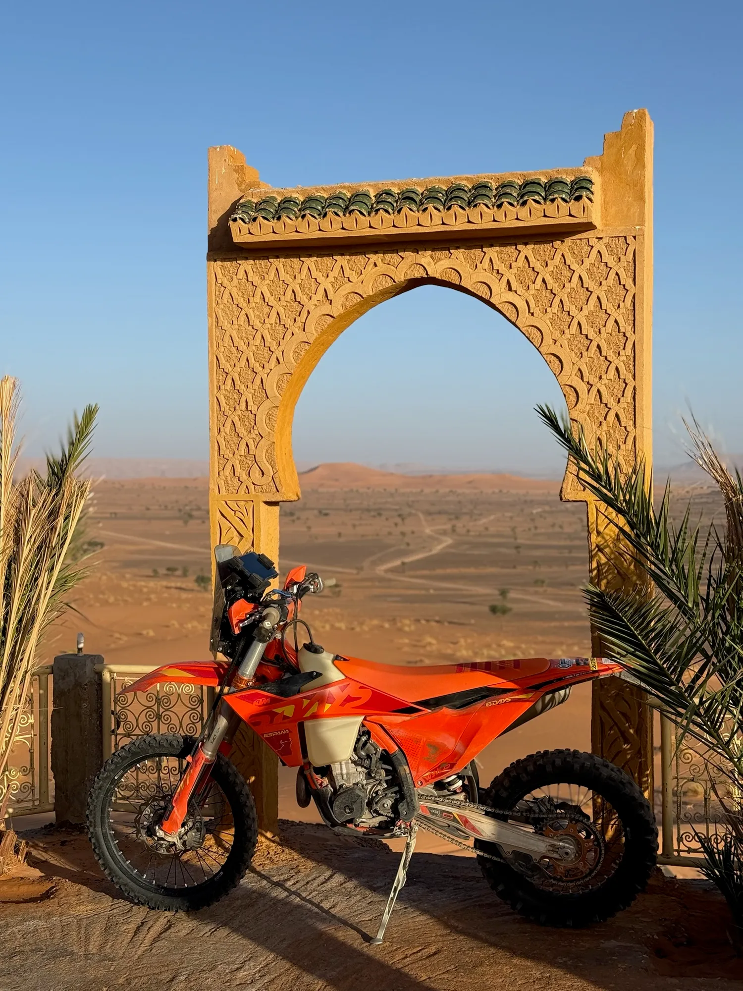 KTM sous une arche marocaine ornée, dunes en arrière-plan au couchant