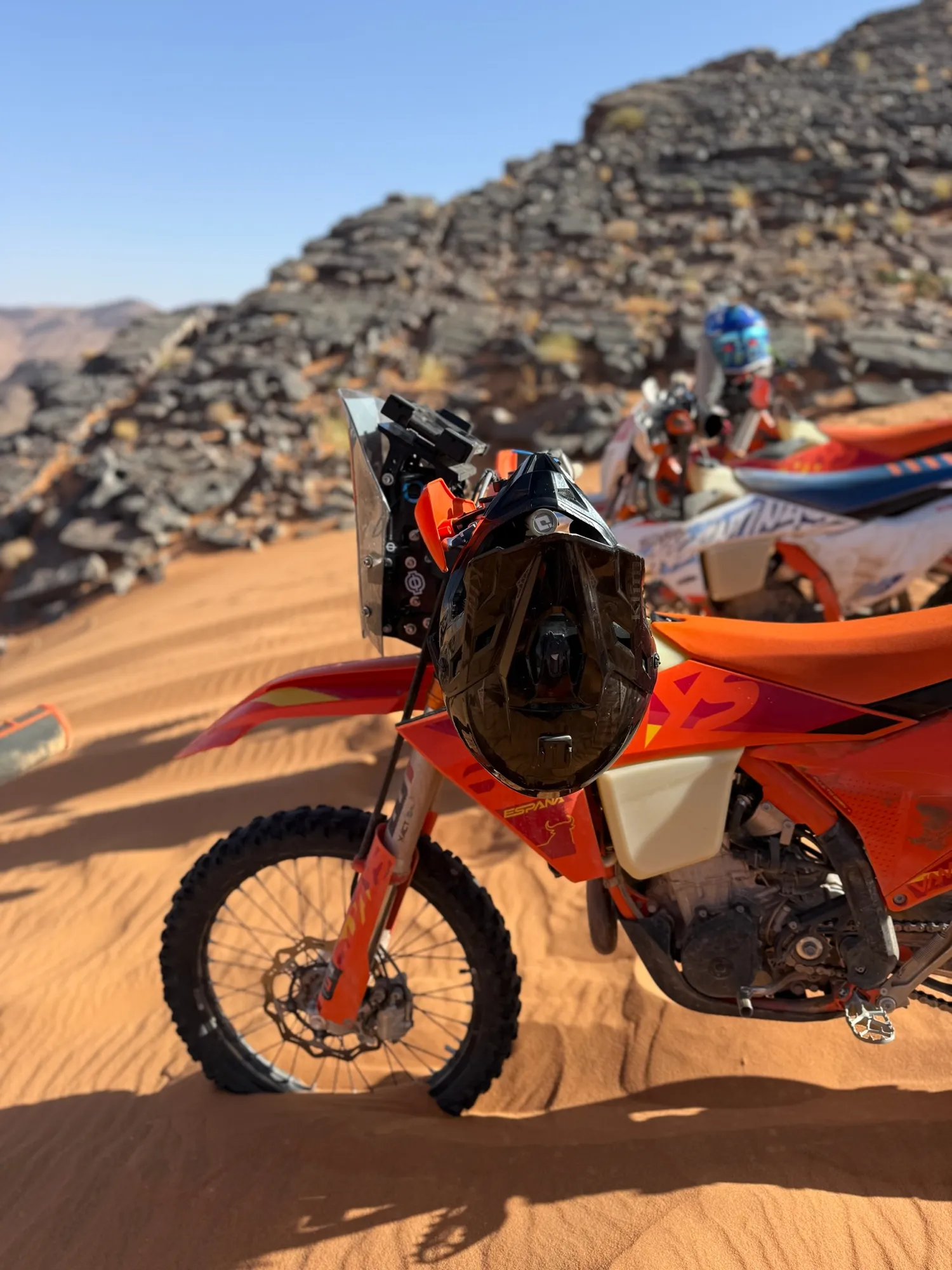KTM et casque posés sur le sable, rochers noirs en fond